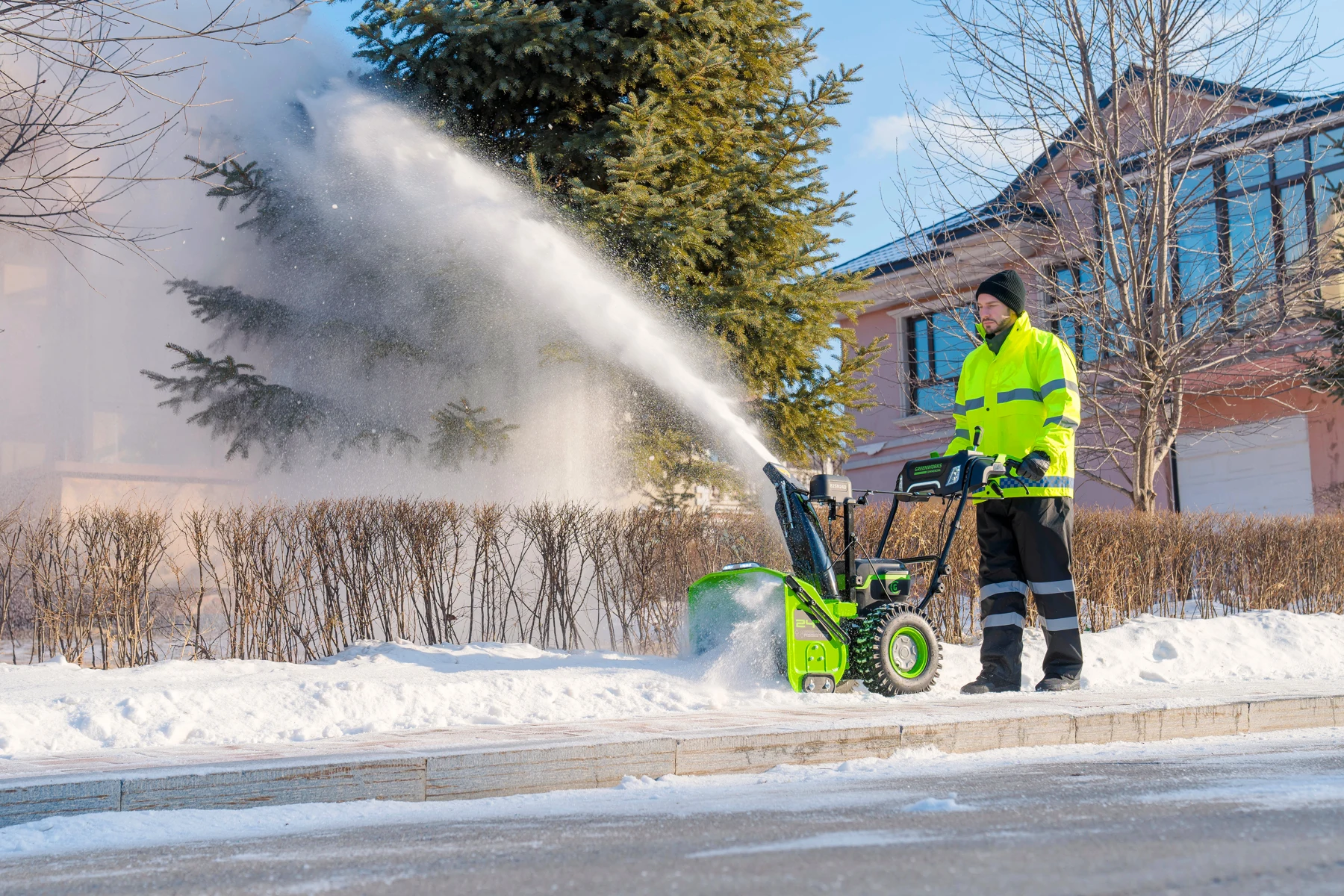 Зимнее утро: человек расчищает снег у дома снегоуборщиком Greenworks